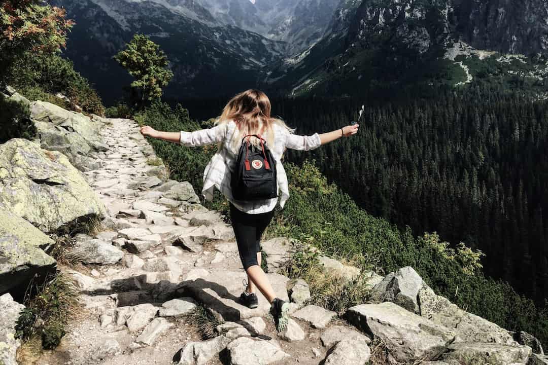 Woman with a black backpack walking on a rocky mountain trail, arms stretched out, surrounded by green forest and rugged peaks