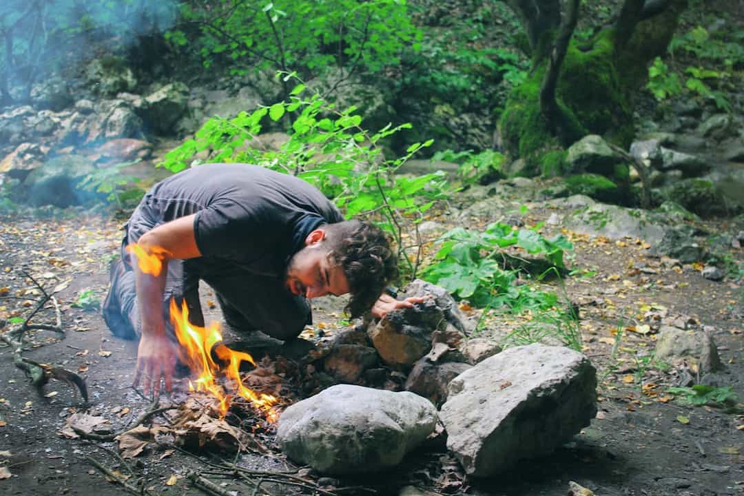 Man crouched in a forest clearing, tending to a small campfire surrounded by rocks and dry leaves