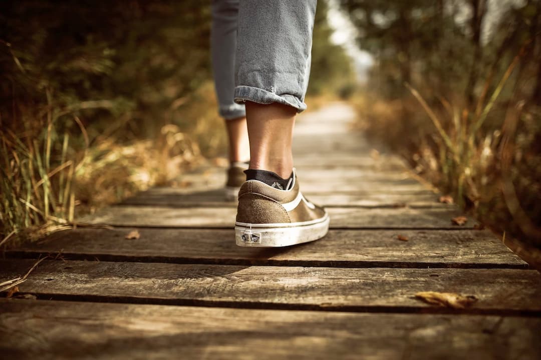 Close-up of a person wearing rolled-up jeans and brown Vans sneakers walking on a wooden boardwalk surrounded by grass and trees