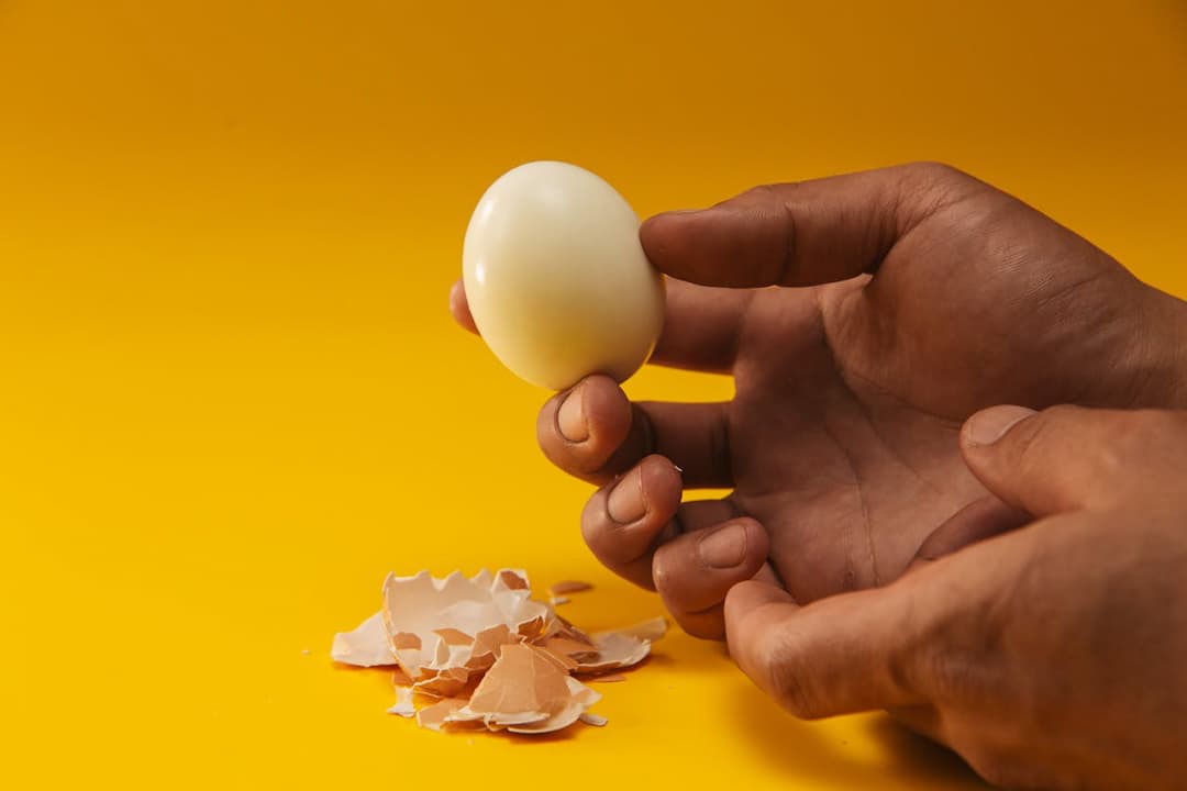 Person holding a peeled boiled egg over a bright yellow background, egg shell fragments scattered on the surface below