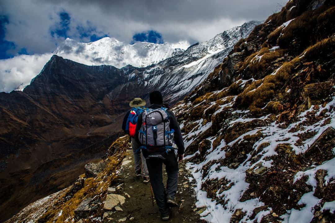 Two hikers with backpacks walking on a narrow snowy mountain trail, surrounded by rugged peaks and dramatic clouds