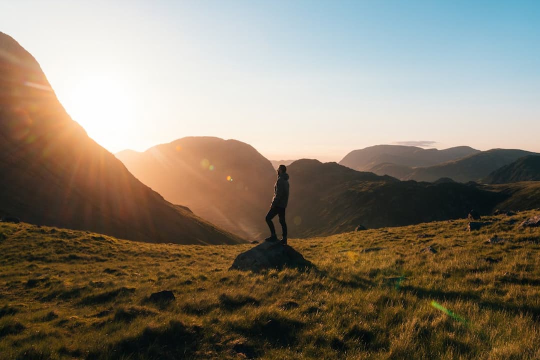 Person standing on a rock in grassy valley, facing mountain range during sunrise, golden light casting long shadows