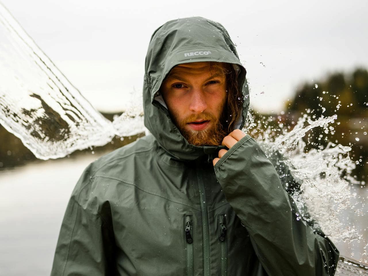 Man in green RECCO waterproof jacket with hood up as water splashes off, outdoors near a body of water