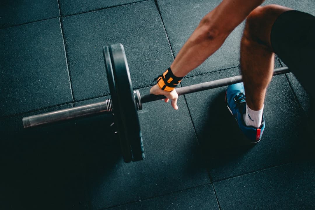 Person gripping a barbell with weight plates in a gym, wearing an orange wrist strap, blue sneakers, and standing on black rubber flooring