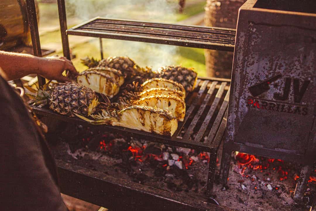 Grilled pineapple halves on an outdoor barbecue over glowing charcoal, hand adjusting one piece