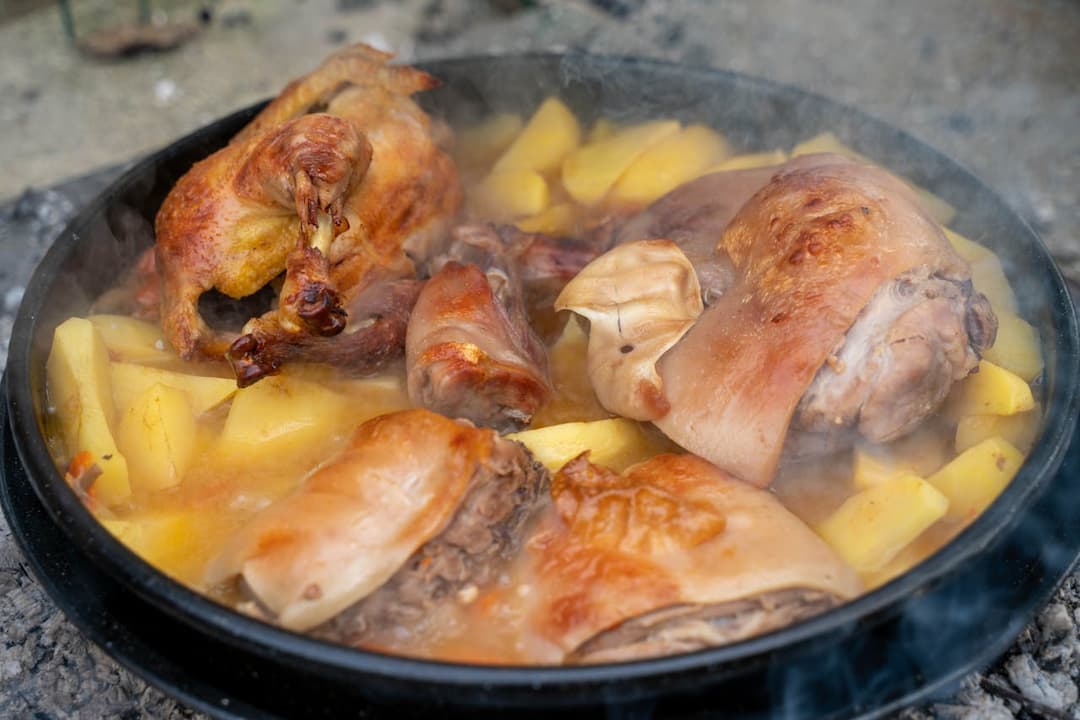 teaming hot stew of roasted chicken, pork, and sliced potatoes in a black pan over an open fire