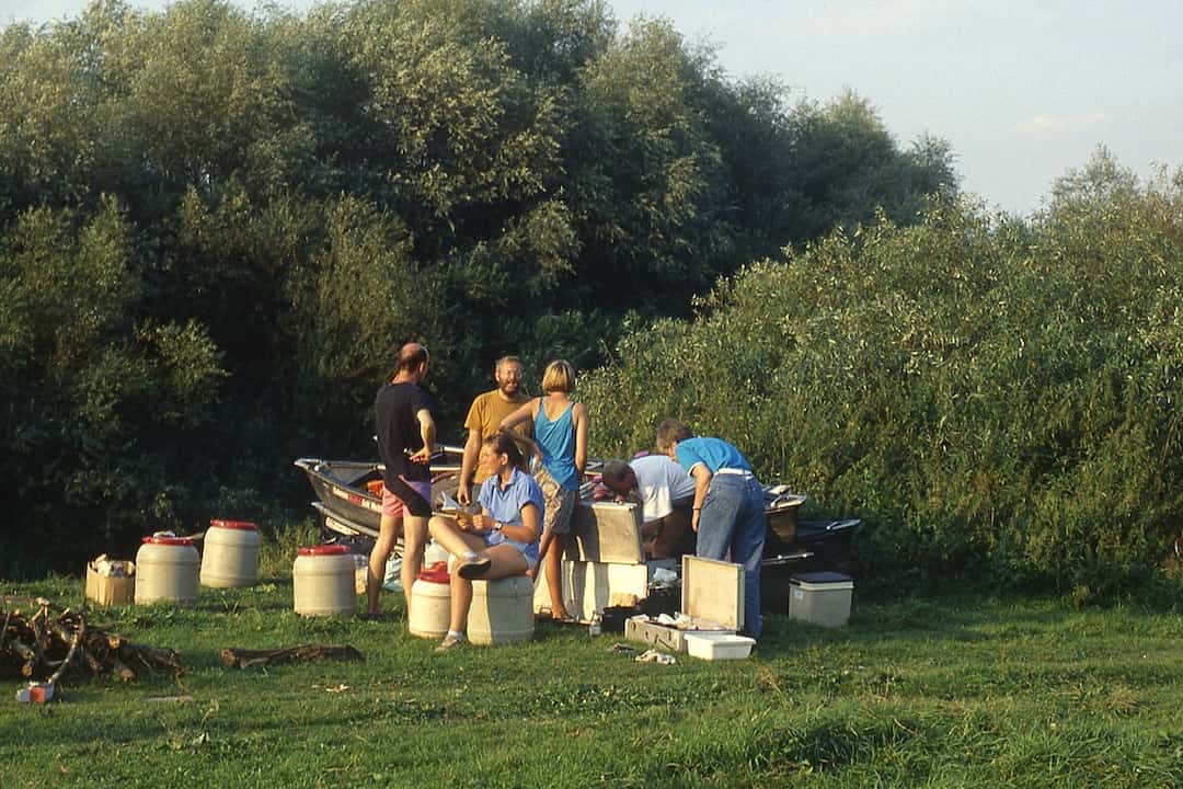 Group of people at a grassy campsite, some seated on barrels and others preparing items around a boat and gear, with dense green trees in the background