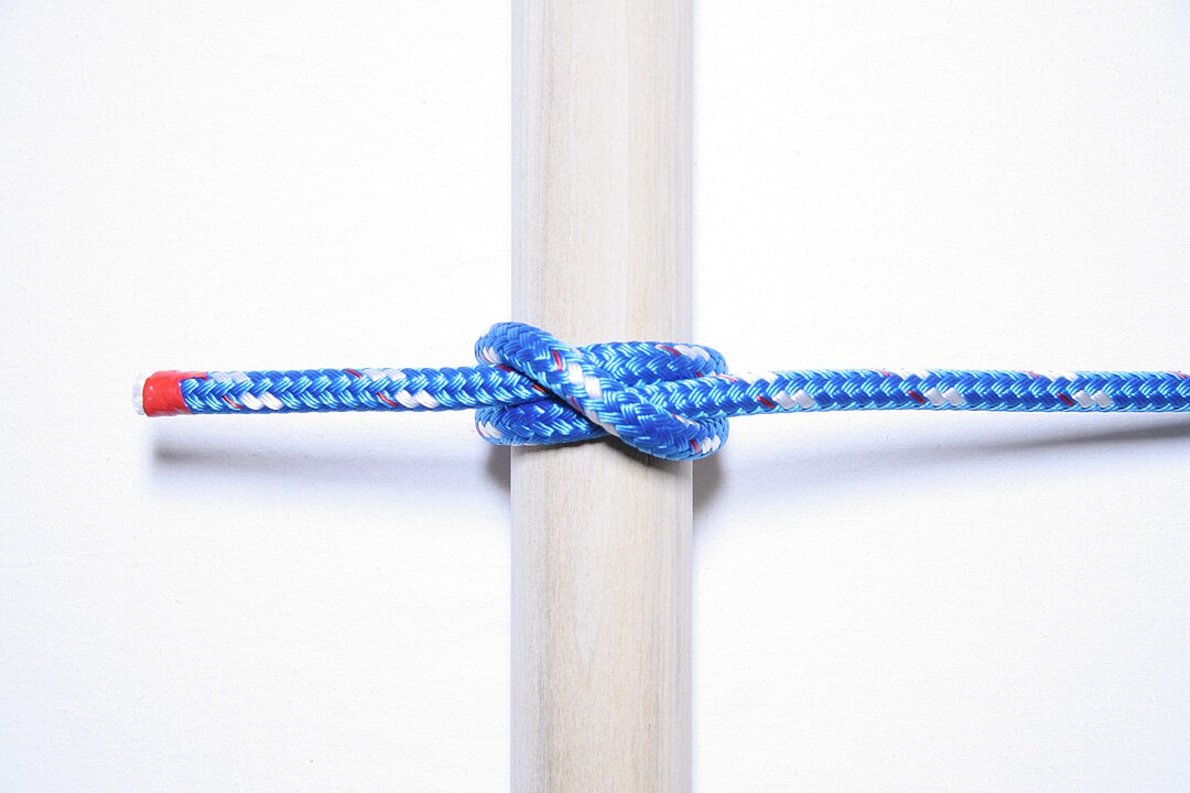 A clove hitch knot tied with a blue, red, and white braided rope around a vertical wooden pole on a white background