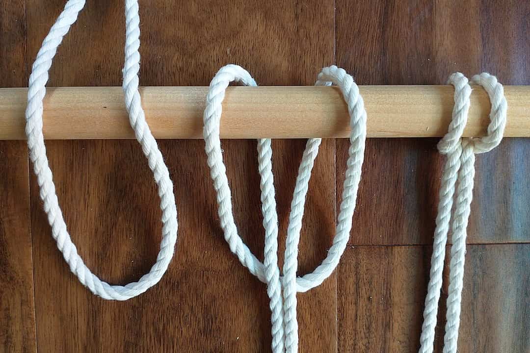 Three white ropes tied in different macramé anchor knots on a horizontal wooden dowel over a dark wooden backgroun