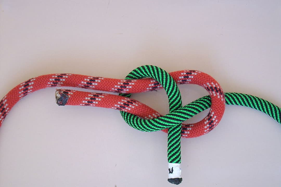A sheet bend knot joining a red rope with white and black patterns to a green and black striped rope against a light background