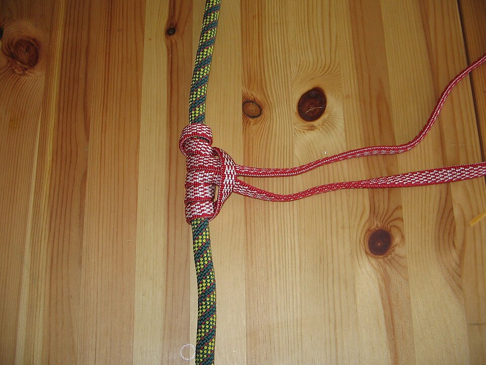 Red cord tied in a three-turn Prusik knot around a green climbing rope on a wooden surface