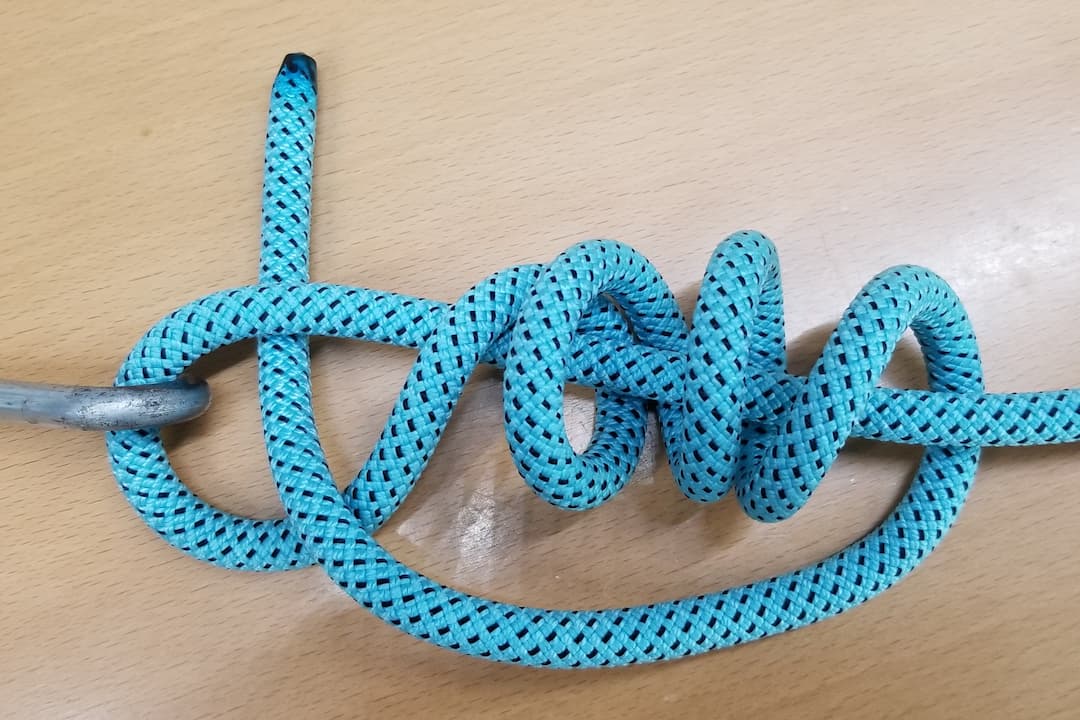 Blue rope tied in a Clinch Knot around a metal hook, resting on a wooden surface, with multiple tight coils and the tag end looped back through the coils