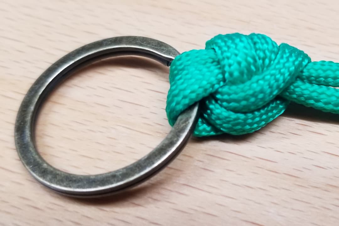Close-up of a green braided cord tied to a metal ring using a Palomar knot, placed on a light wooden surface