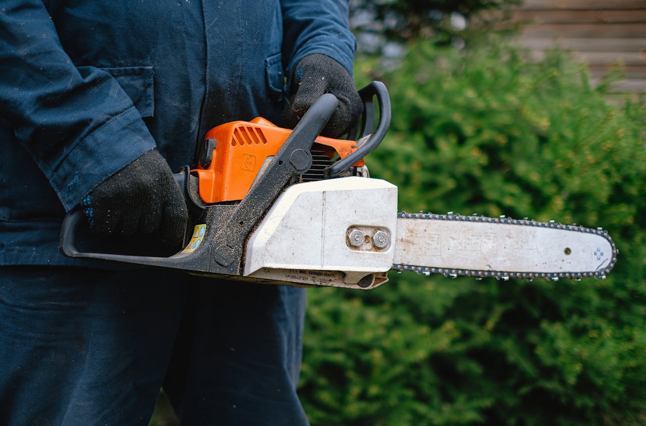 erson in dark workwear holding a gas-powered chainsaw with gloves, standing in front of green bushes
