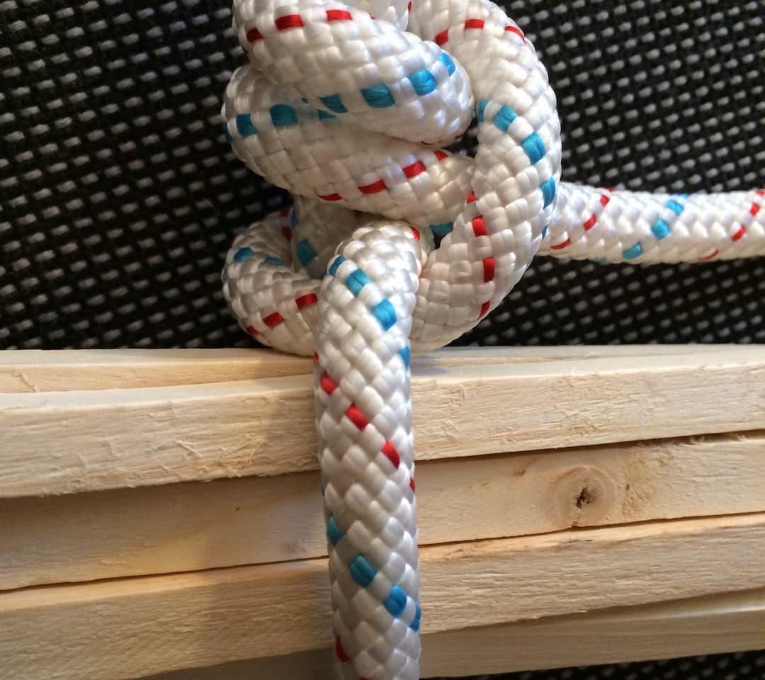 White braided rope with red and blue tracers tied in a San Diego Jam Knot around stacked wooden slats, placed in front of a black textured surface