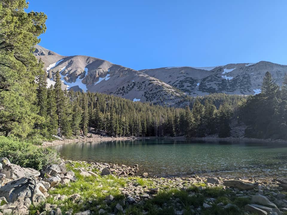 Alpine lake surrounded by pine forest with snow-dusted rocky mountains in the background, under a clear blue sky at Great Basin National Park