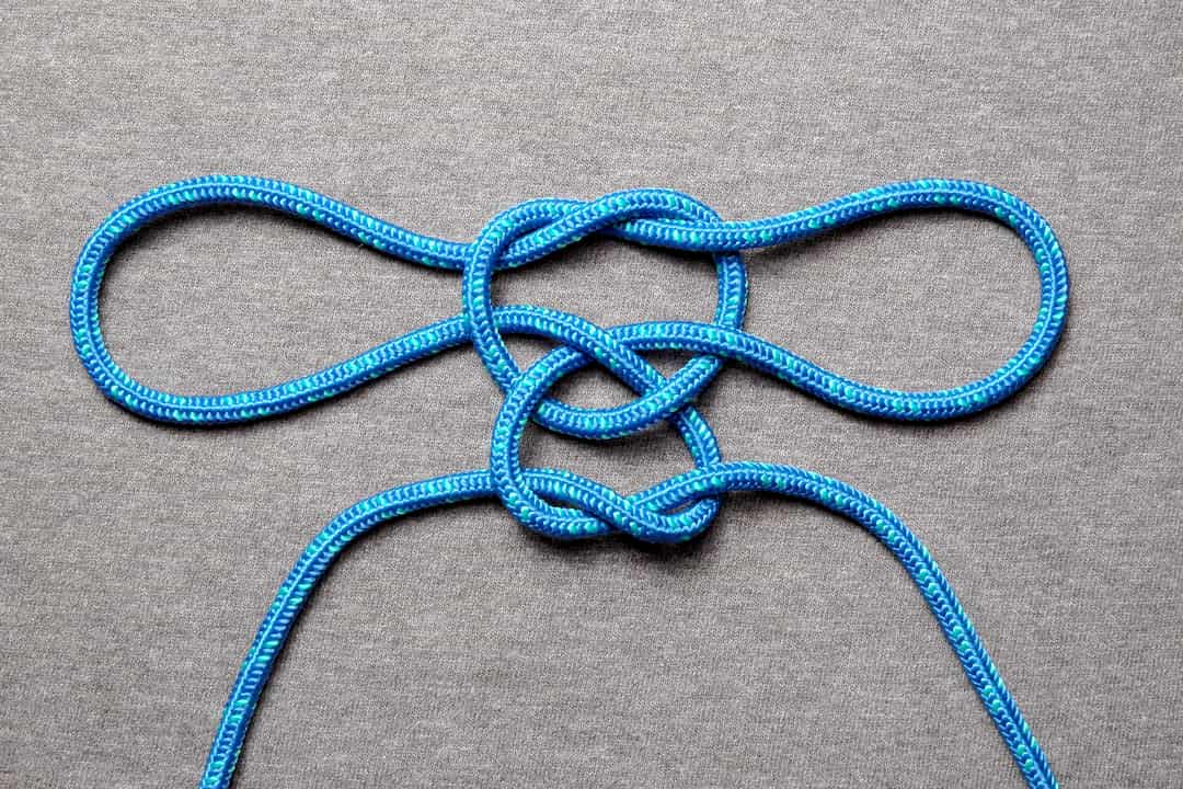 Blue rope tied in a reef knot variation resembling a handcuff knot, with two symmetrical loops on either side and a central crossing, laid flat on a grey fabric surface