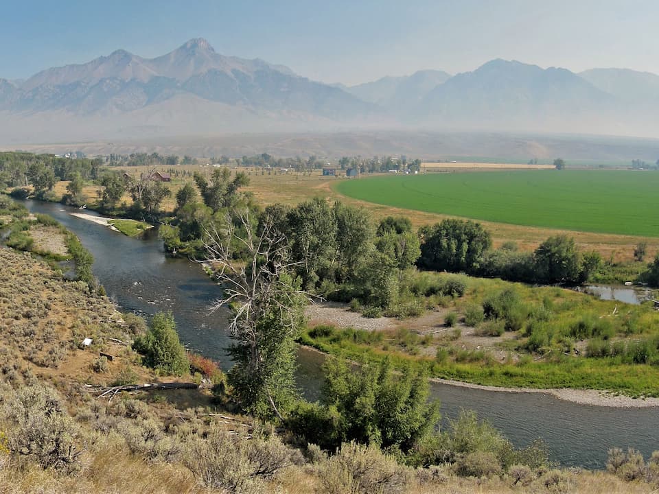 Winding river bordered by green trees and dry brushland, flowing through a wide valley with irrigated farmland