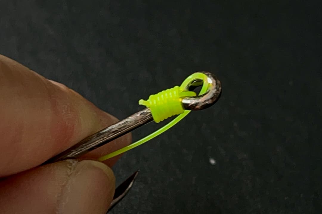 Snell Knot tied with bright yellow fishing line onto the eye of a fishing hook, held between fingers against a dark background