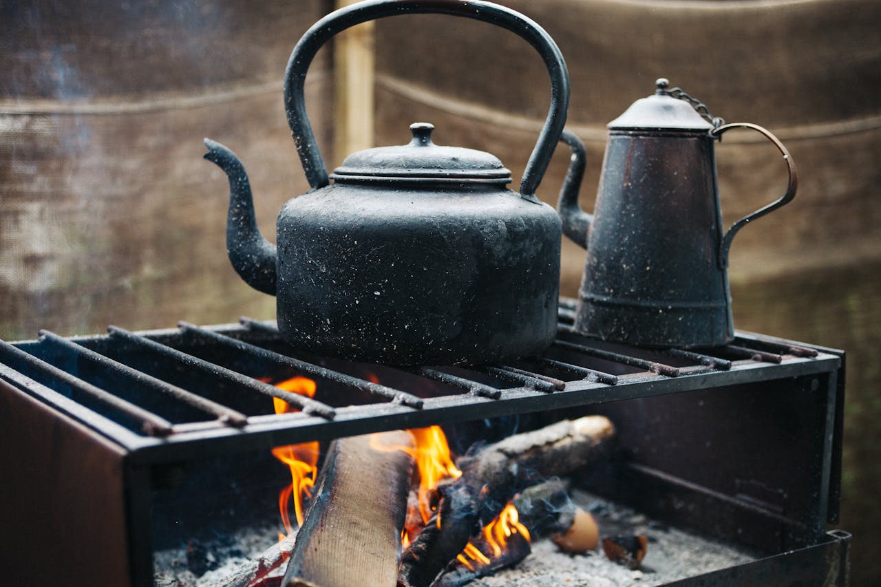 Blackened kettle and metal pitcher on a woodstove grill over an open flame, with logs burning beneath