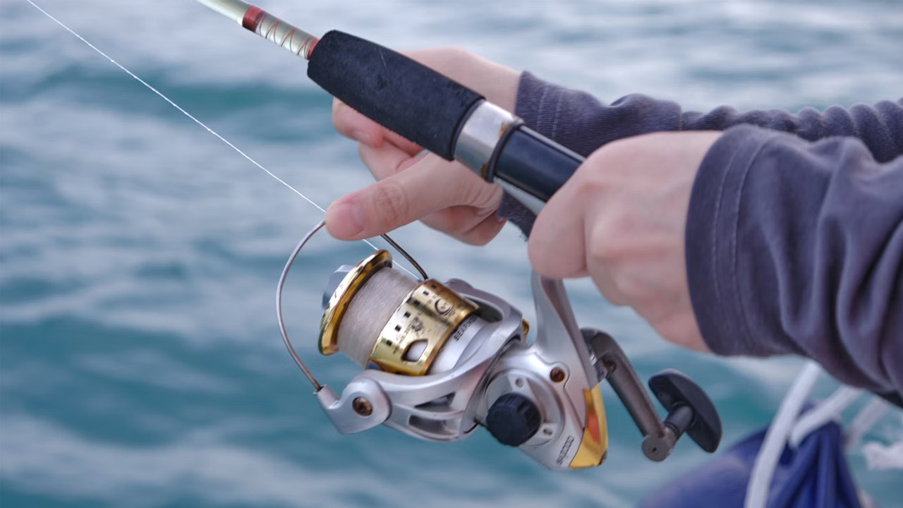 Close-up of hands holding fishing rod, spinning reel with gold and silver accents, person wearing long-sleeved shirt, calm blue water in background