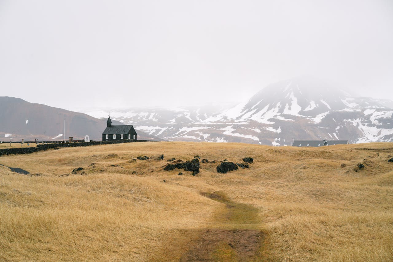 Small black church in a foggy, snow-capped mountain landscape with golden grass and volcanic rocks in the foreground