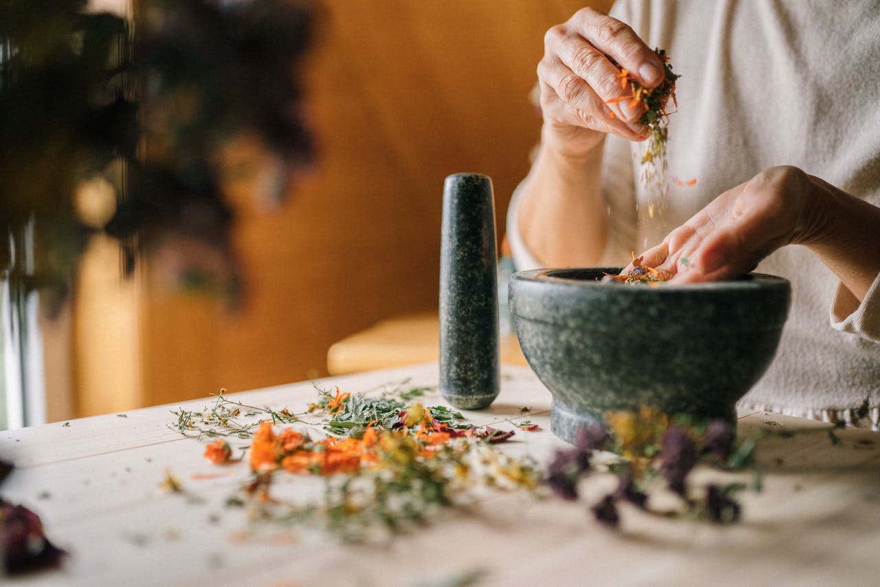 Person preparing dried medicinal herbs in a stone mortar with pestle on a wooden table.