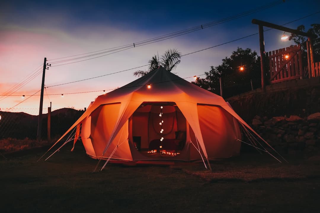 Large glamping-style tent illuminated from within, set up outdoors at dusk with string lights and cozy interior decor