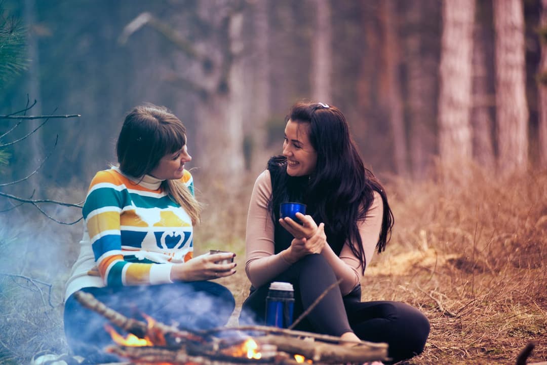 Two women sitting by a campfire in the woods, smiling and holding cups, with a thermos nearby