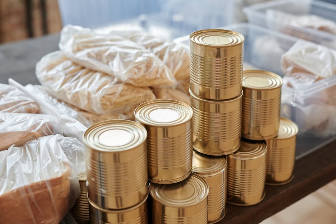 Stack of golden tin cans and packaged bread and pasta on a wooden table, prepared for food distribution
