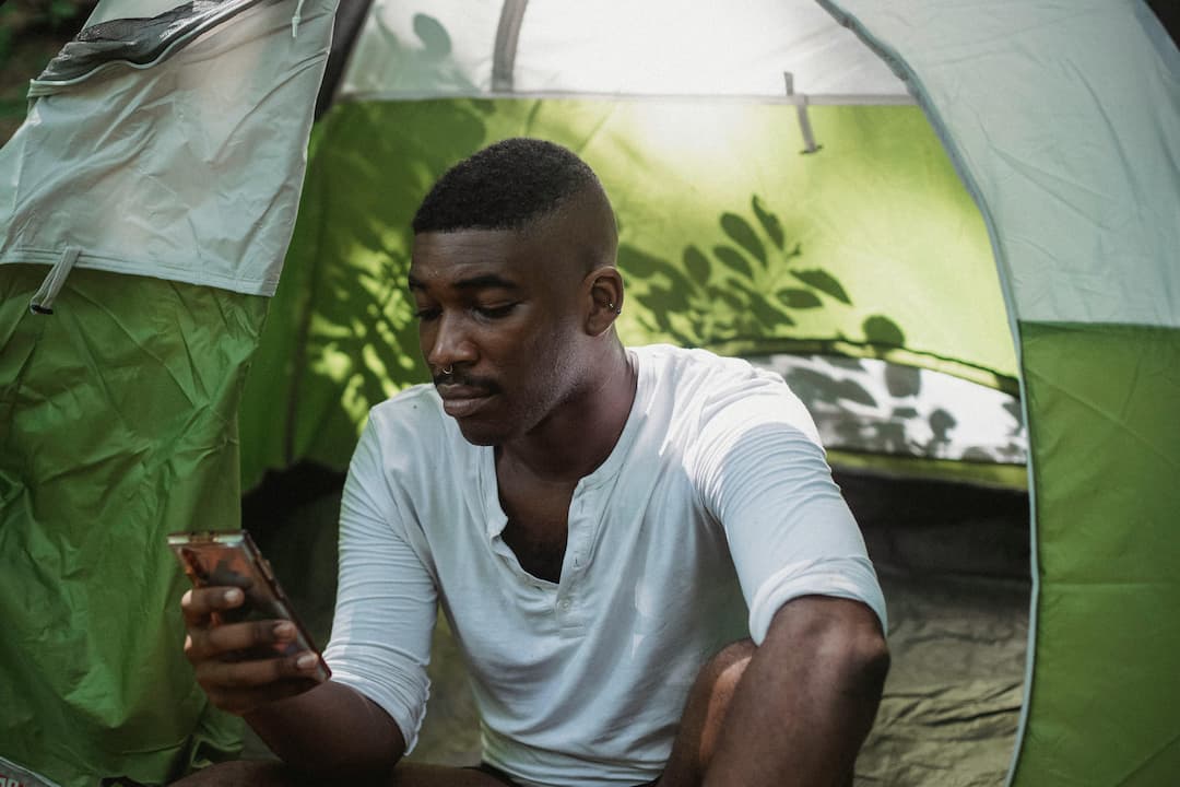 Black man in a white shirt sitting at the entrance of a green tent, looking at his smartphone