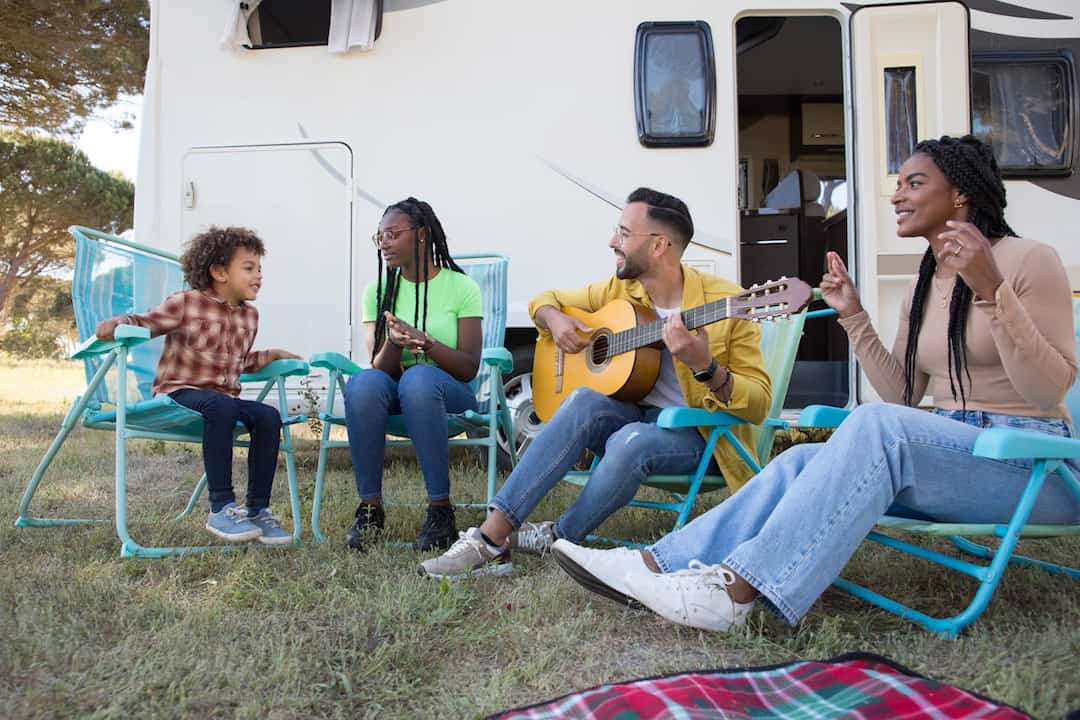 Group of four adults and one child sitting on camping chairs outside a camper van, enjoying music and clapping along to a guitar player
