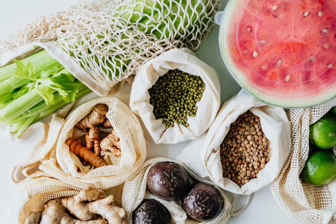 Assorted fresh produce including celery, ginger, turmeric, limes, green mung beans, brown lentils, purple passion fruits, and sliced watermelon, stored in reusable mesh and cloth bags on a white surface