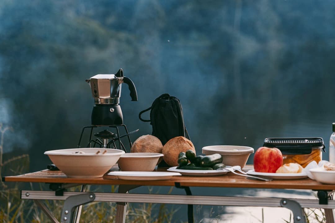 Camping table setup near water, featuring a moka pot on a portable stove, coconuts, bowls, an apple, pickled vegetables, cucumbers, bottled water, and assorted food items