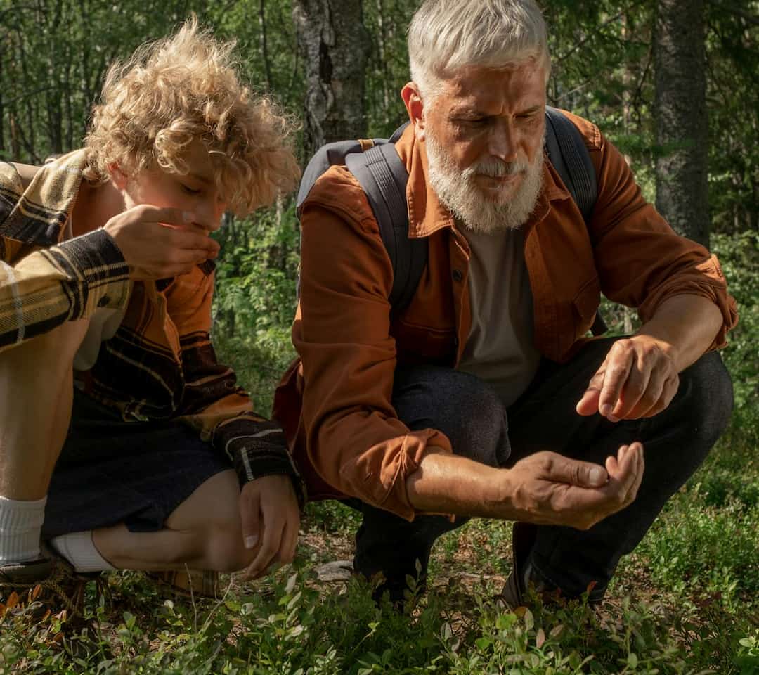 Two people crouched in a forest clearing, one inspecting a plant closely in his hand, the other appearing to taste or smell a gathered item