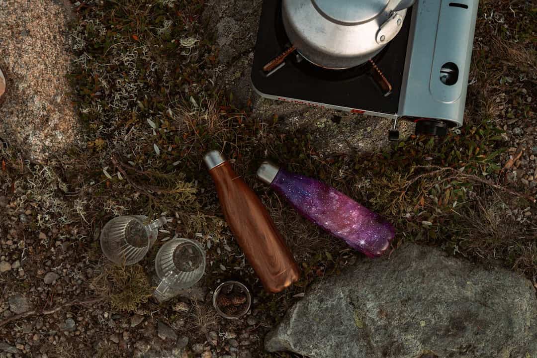 Outdoor camping scene, portable gas stove with a kettle, wooden and galaxy-patterned insulated bottles, two glass mugs, rocky ground with moss and plants