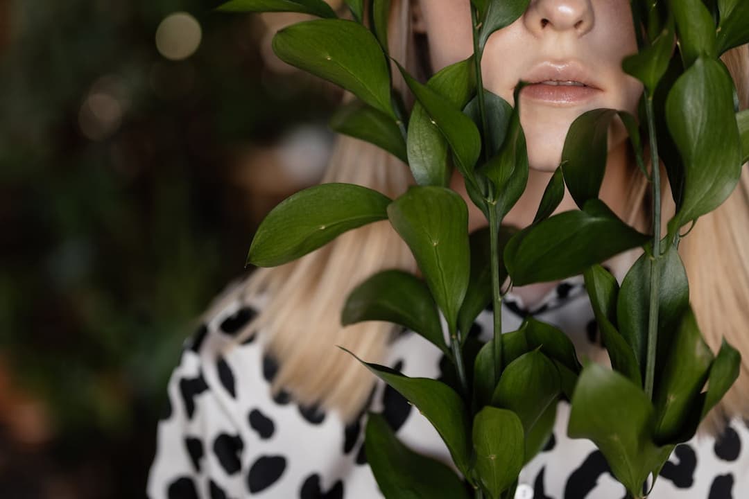 Woman with blonde hair and cow-print shirt partially hidden behind lush green leaves, plant stem appearing close to her lips