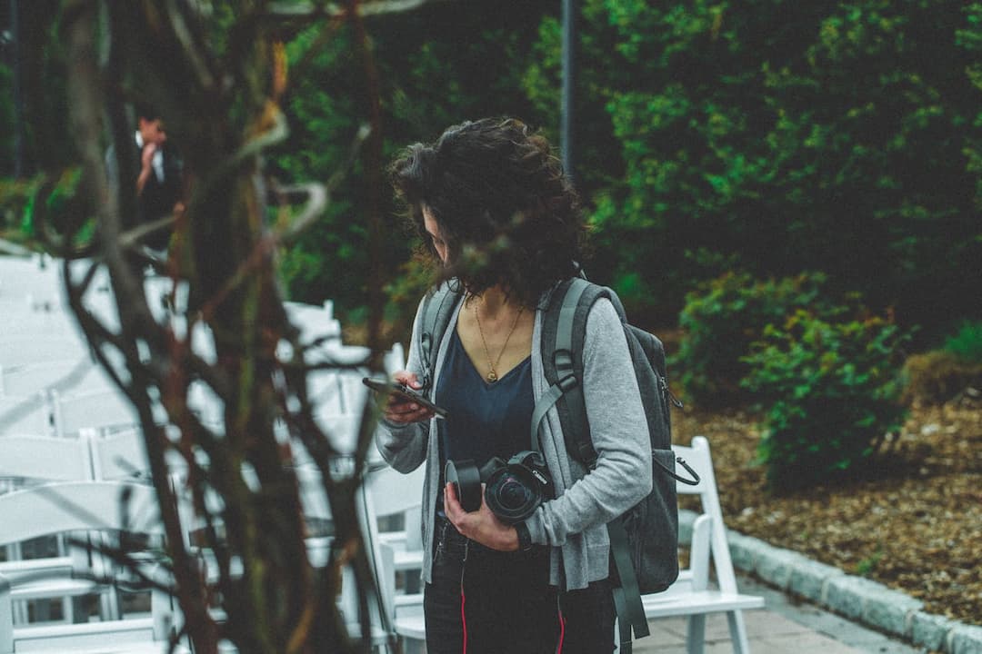 Woman holding a camera and phone, wearing a backpack, standing near rows of white chairs in an outdoor setting