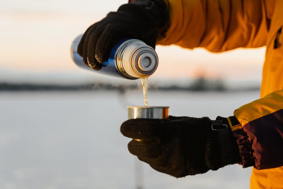 Person in winter gloves and yellow jacket pouring hot liquid from a thermos into a metal cup outdoors in a snowy landscape