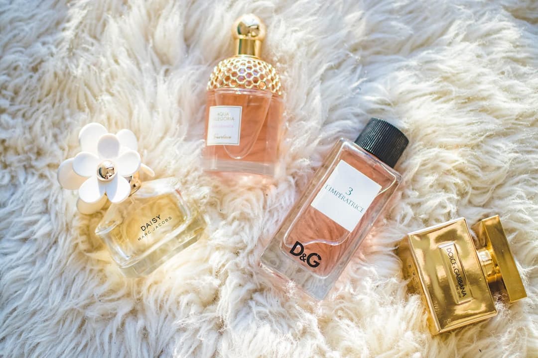 Four luxury perfume bottles, including Daisy by Marc Jacobs, Aqua Allegoria by Guerlain, 3 L’Imperatrice by D&G, and a gold Dolce & Gabbana bottle, arranged on soft white fur