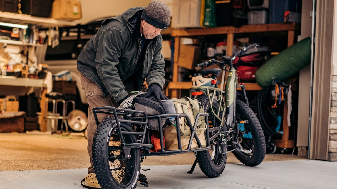 Man packing gear onto cargo electric bike, wearing black beanie and outdoor clothing, parked in front of open garage with tools and equipment