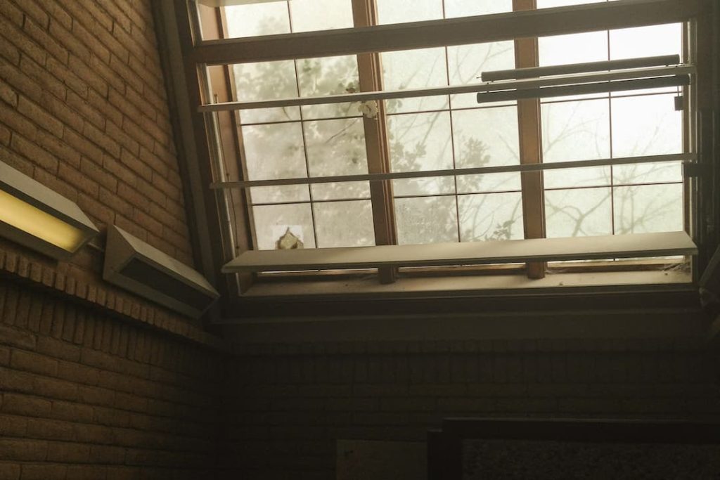 Interior view of a slanted skylight window with frosted glass and wooden framing, mounted on a brick wall, with visible fluorescent light fixtures on the side