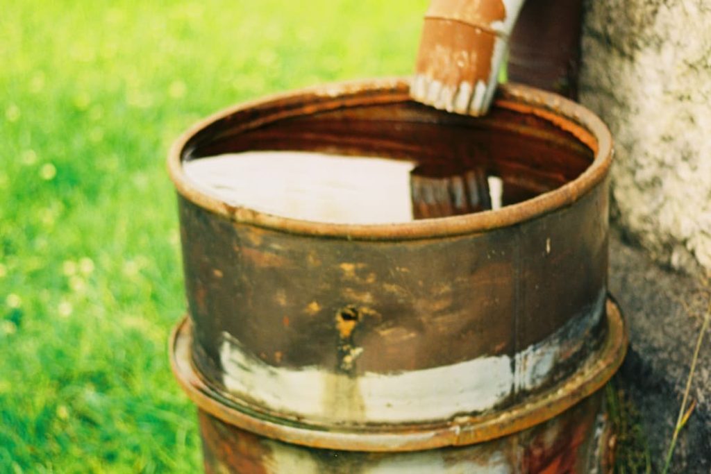 Rusty metal barrel collecting rainwater from a brown downspout, positioned beside a stone wall on a grassy yard