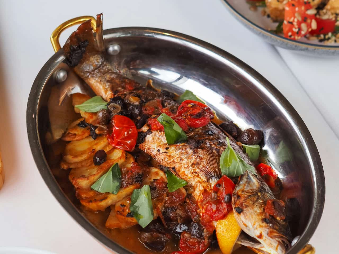 Steamed whole mountain trout served in a metal dish with potatoes, cherry tomatoes, olives, and fresh basil