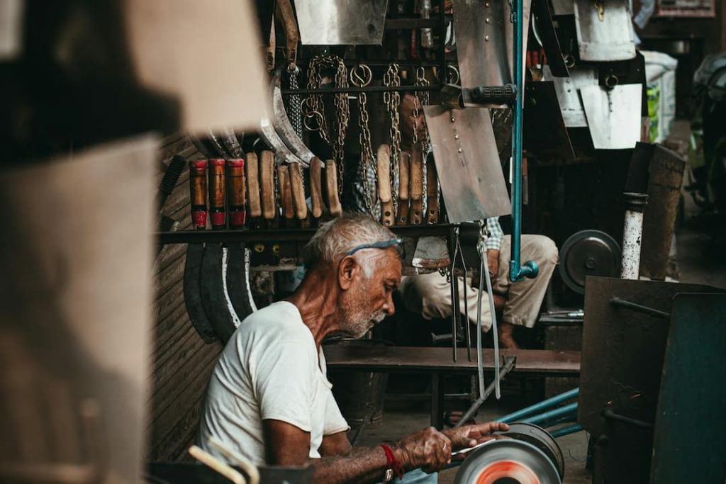 Elderly man sharpening a blade on a grinding wheel in a traditional tool shop surrounded by various knives and metal tools